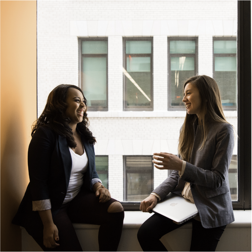two women are sitting on a window sill talking to each other.