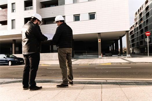 Dos hombres con cascos de seguridad están parados en una acera frente a un edificio.