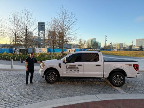 A man stands next to a white Ford pickup truck featuring business branding, parked on a cobblestone area by a city skyline.