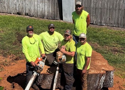 Five people wearing matching neon green shirts and caps pose standing around a large, flat tree stump on a grassy field.