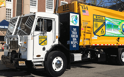 A yellow residential side-loader garbage truck parked on a gravel lot in front of a tree line.