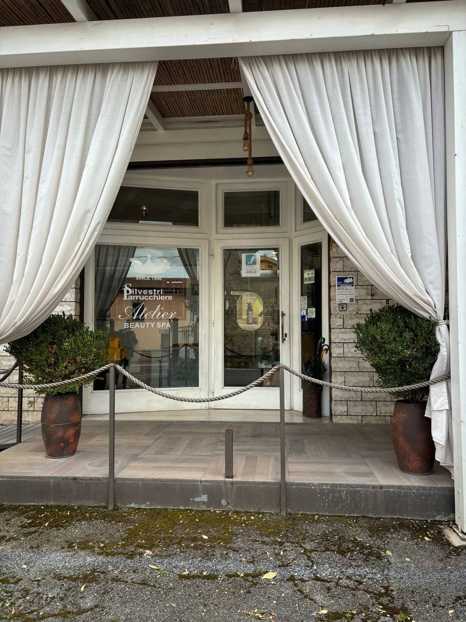 Ingresso del negozio con tende bianche, porte a vetri e piante in vaso su entrambi i lati.