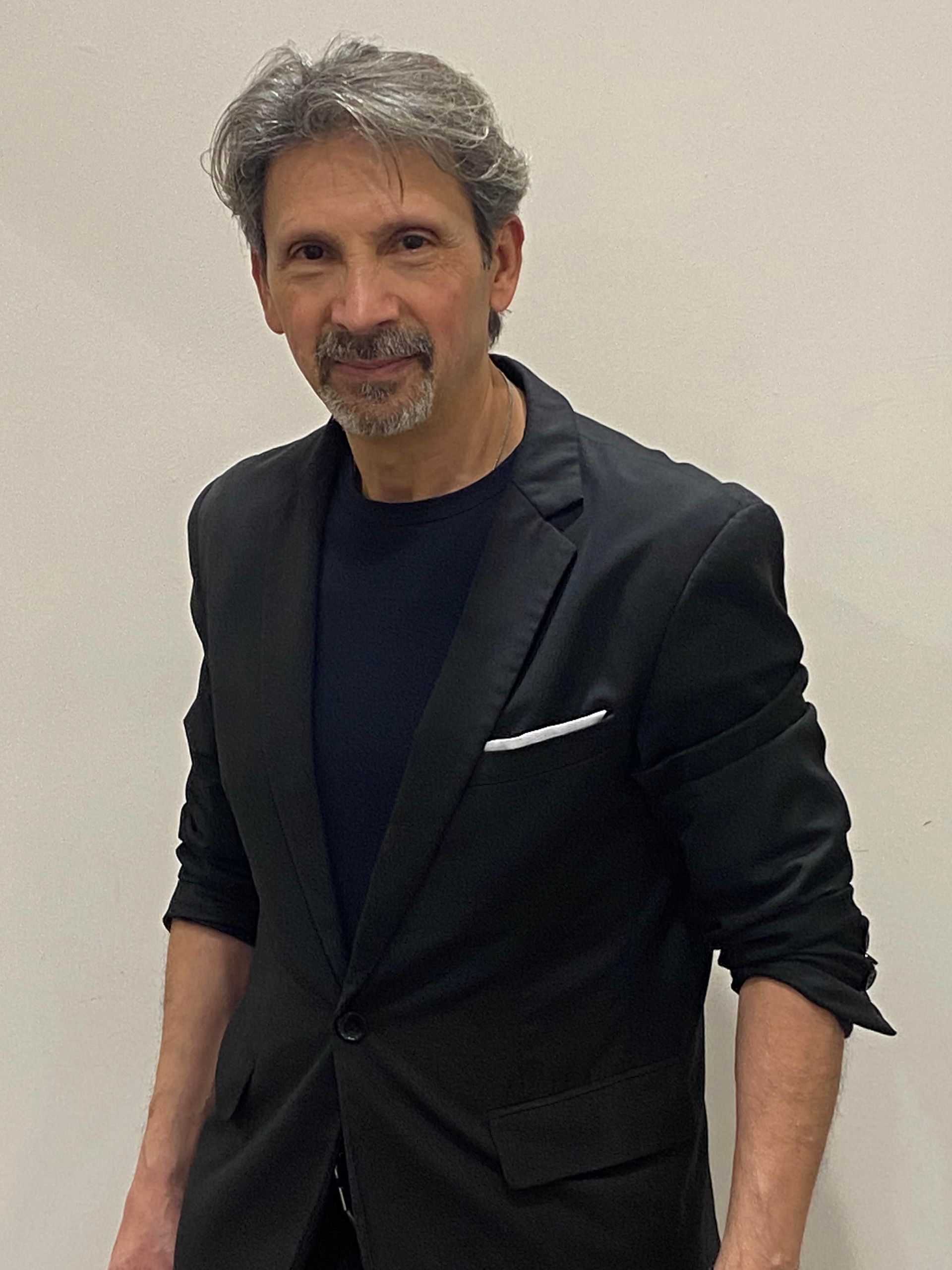 Uomo con blazer nero sopra una camicia scura, in piedi davanti a una parete chiara e sorridente.