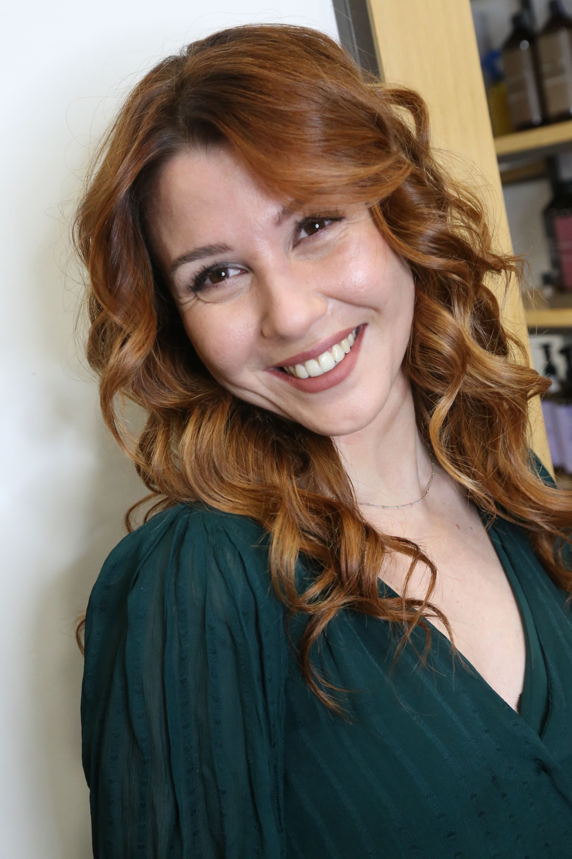 Donna sorridente con lunghi capelli rossi, che indossa una maglietta verde scuro, in piedi in un ambiente interno.