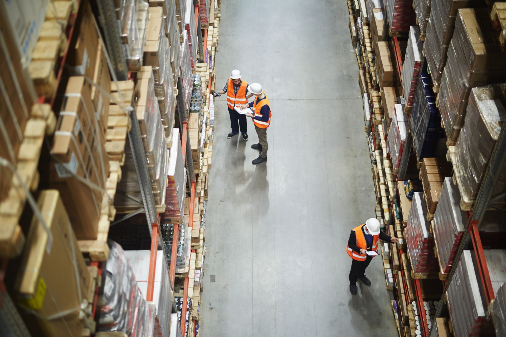 Workers in orange safety vests walk through a warehouse aisle between tall shelves of boxes. Workers in orange safety vests walk through a warehouse aisle between tall shelves of boxes.