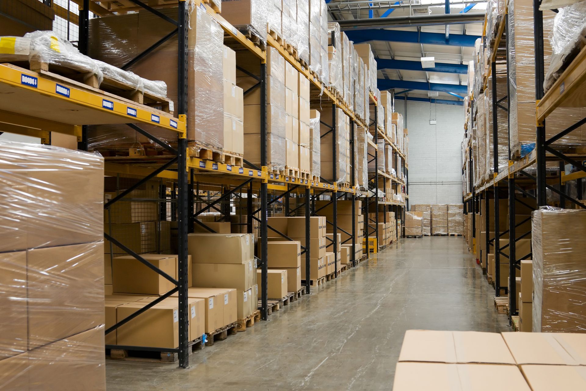 A warehouse aisle with tall shelving, stacked cardboard boxes, and shrink-wrapped pallets. A warehouse aisle with tall shelving, stacked cardboard boxes, and shrink-wrapped pallets.