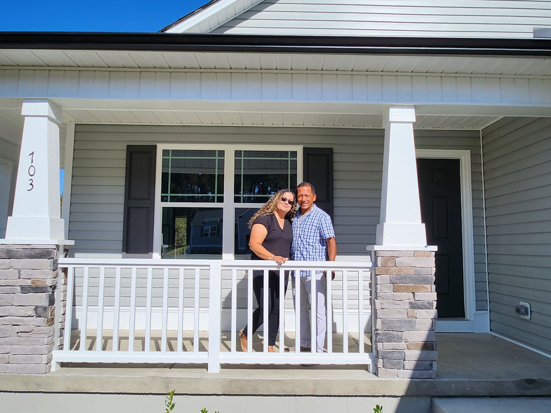 Couple standing on the porch of their new home. Gray siding, white trim, stone accents.