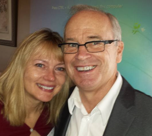 Smiling man and woman posing closely; man in suit and glasses, woman with blonde hair.