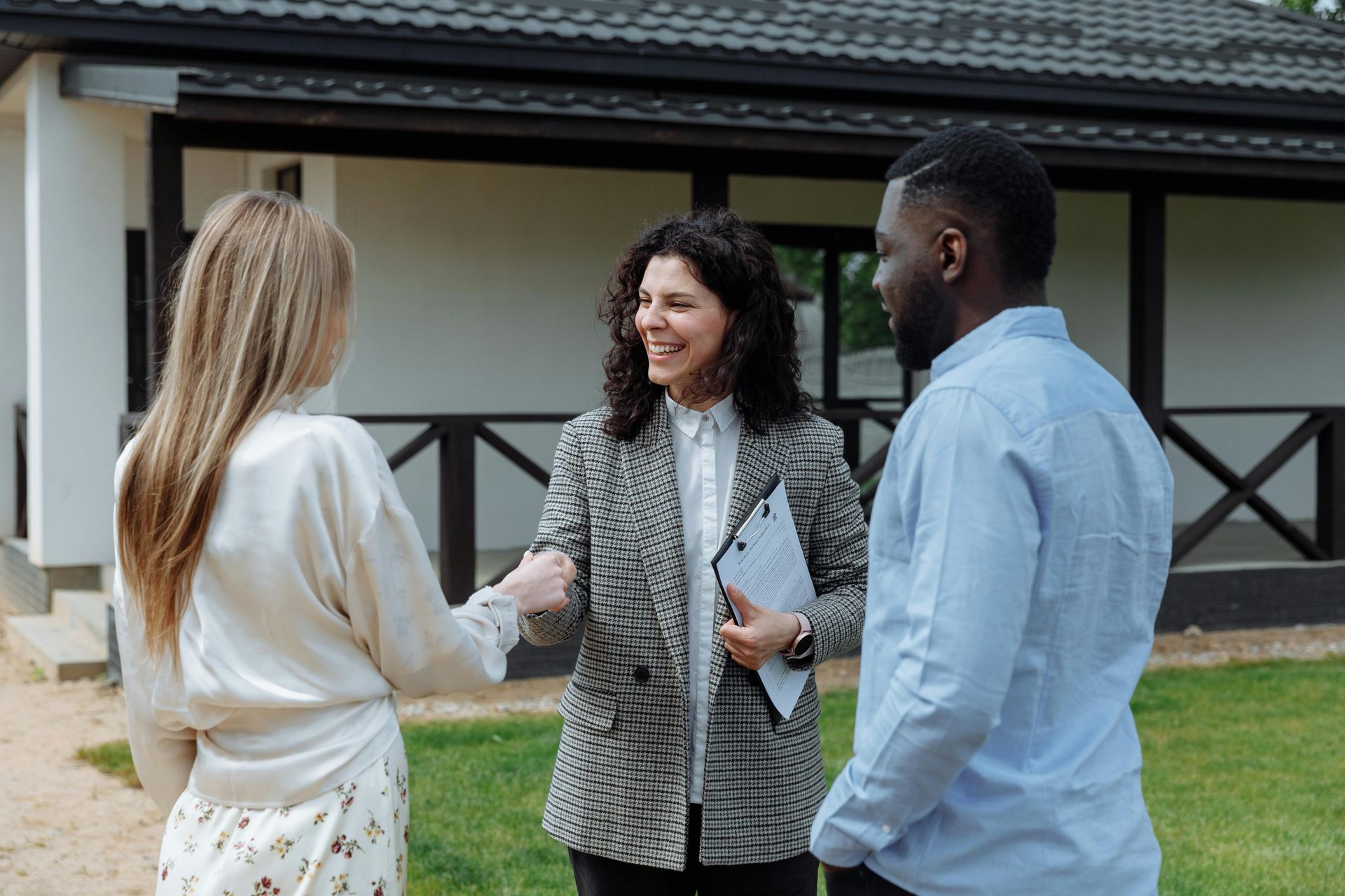 Real estate agent fist-bumps a couple in front of a house; green lawn, sunny day.