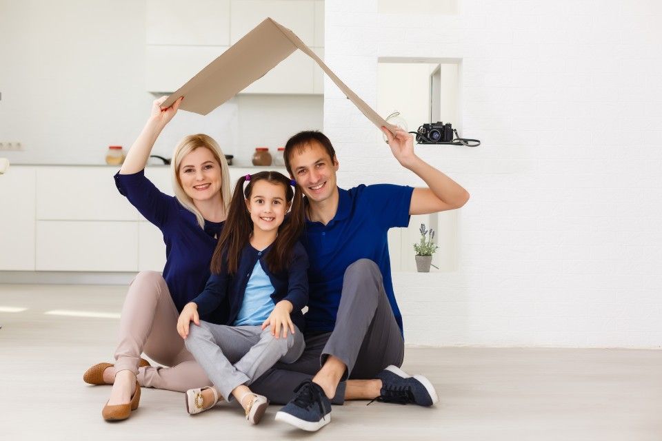 Family of three, sitting under a cardboard roof, smiling, in a bright, modern living room.