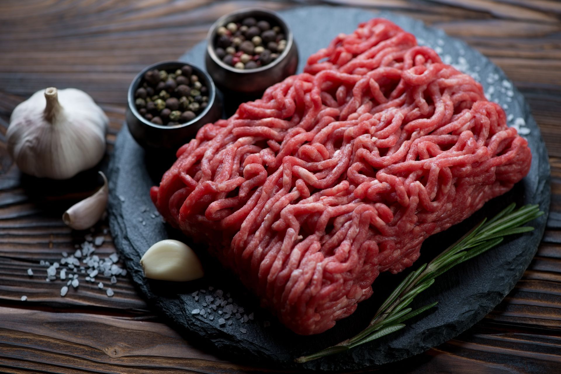 Ground beef on a dark slate with peppercorns, garlic, rosemary, and salt on a wooden surface.