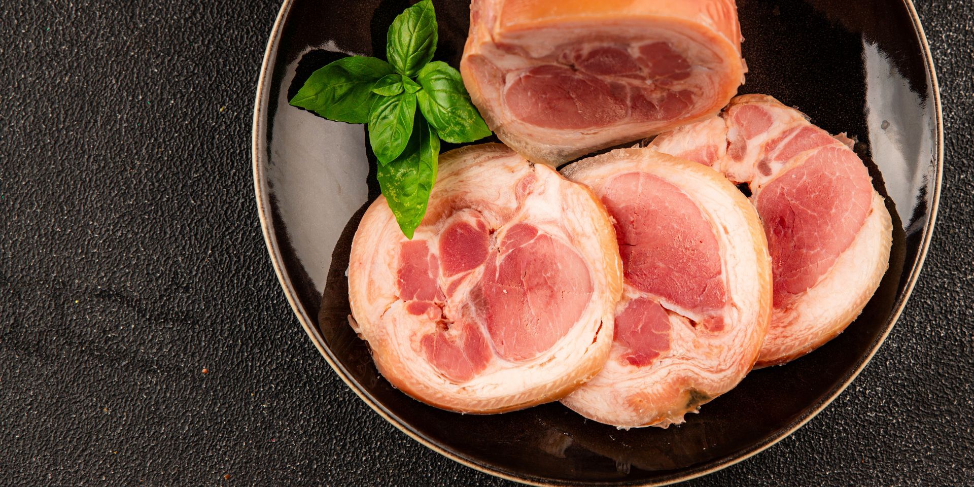 Slices of rolled meat with pink and white marbling, with fresh basil on a black plate, on a dark textured surface.