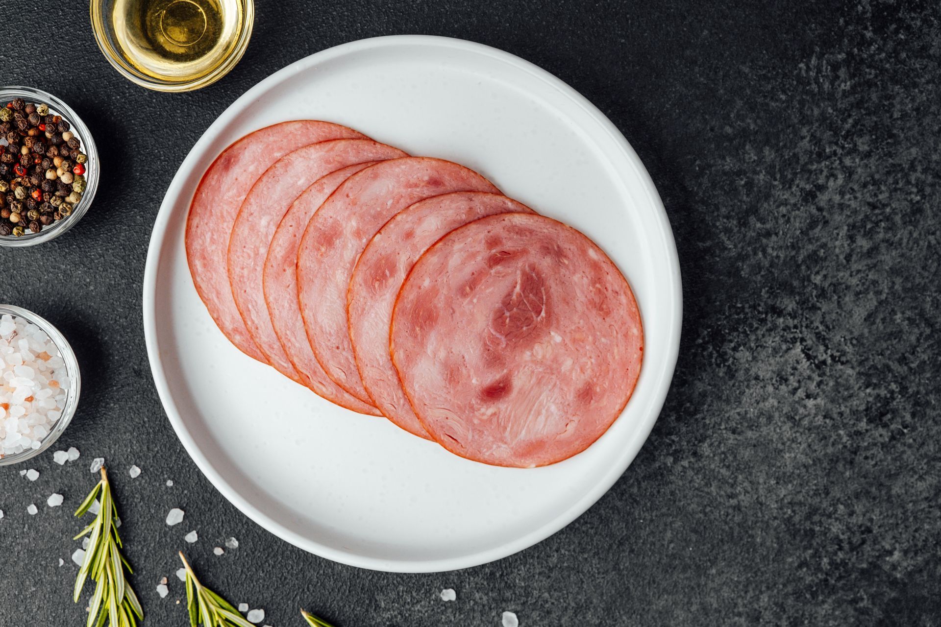 Slices of ham on a white plate, surrounded by spices, oil, and rosemary on a dark surface.