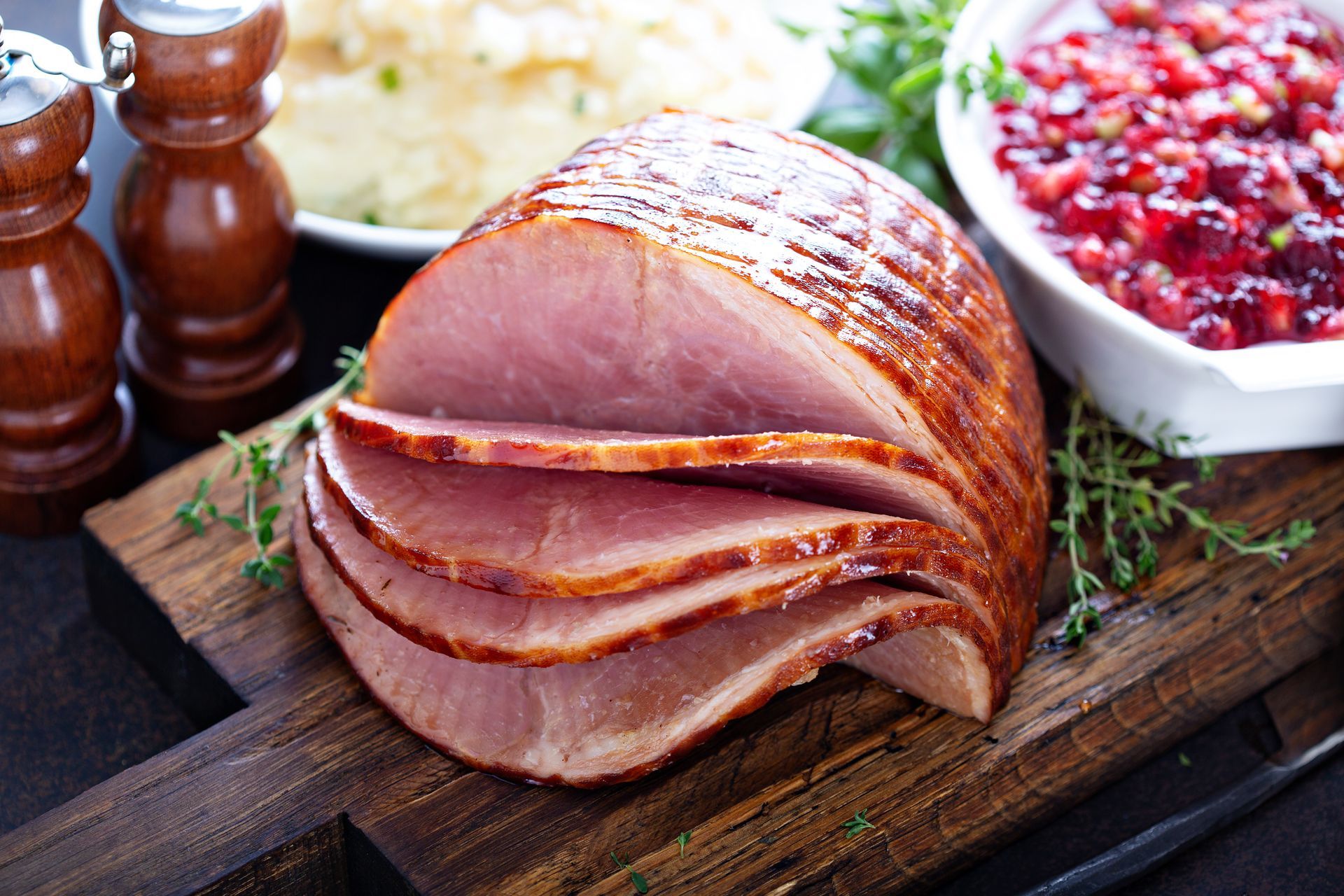 Sliced glazed ham on wooden cutting board, with cranberry sauce and mashed potatoes in background.
