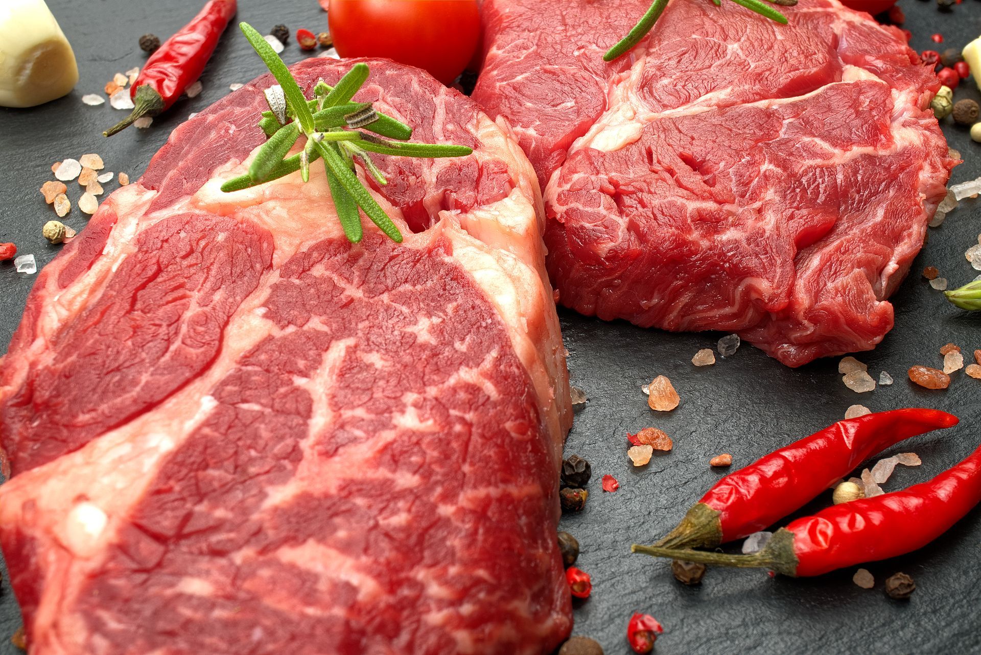 Raw steaks with rosemary sprigs, chili peppers, tomato, and seasonings on a dark surface.