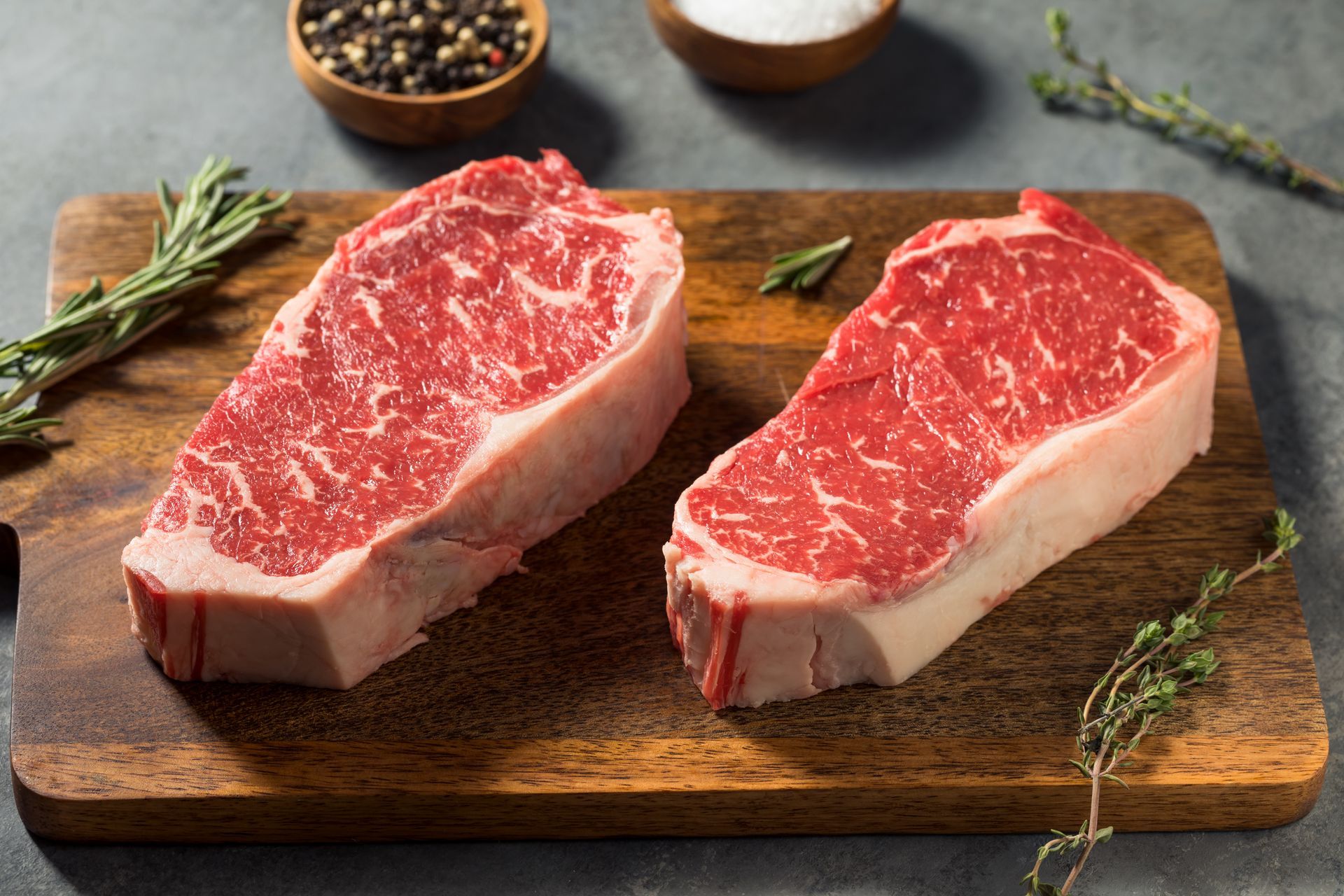 Two raw New York strip steaks on a wooden cutting board with rosemary, salt, and pepper.