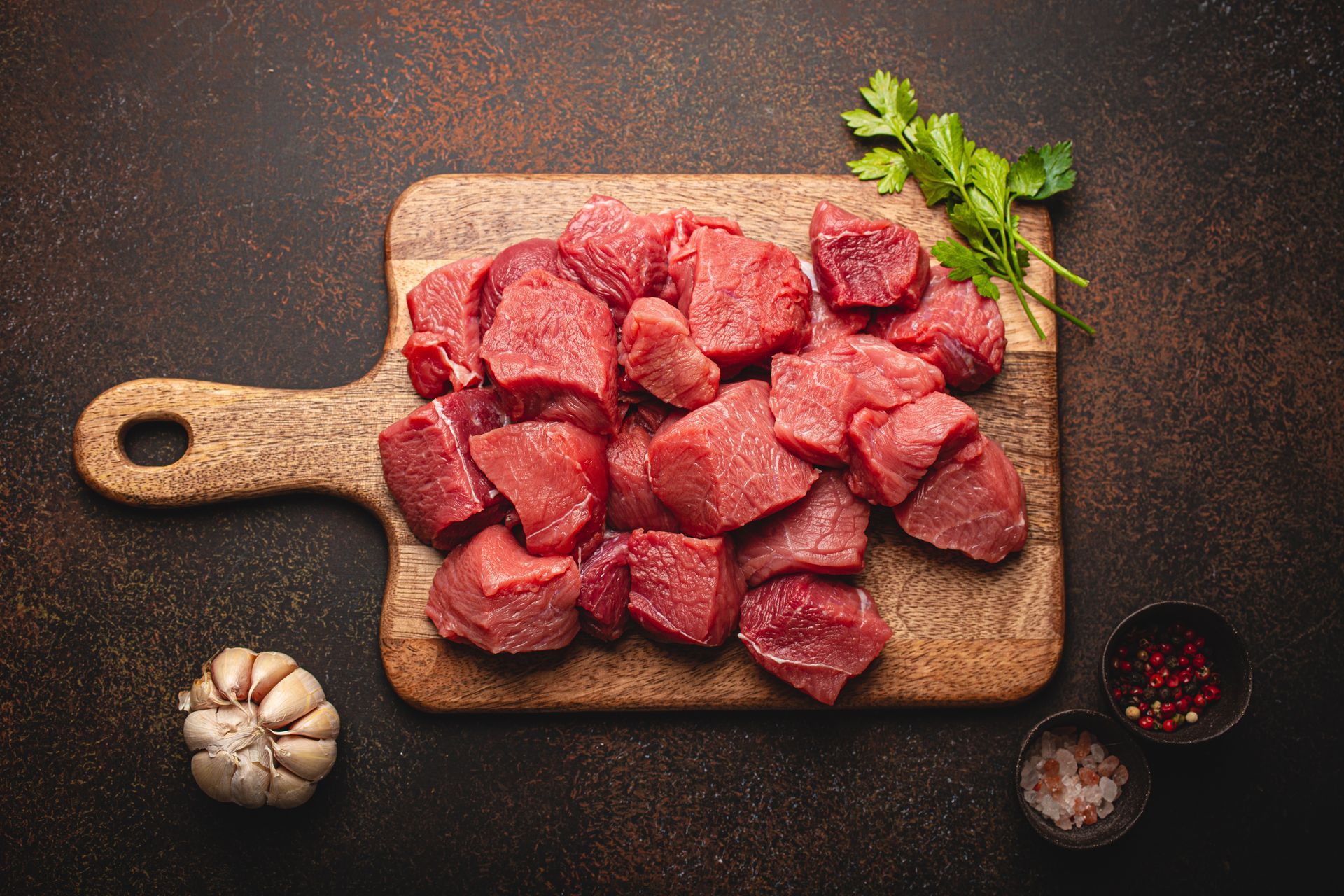 Diced raw beef on a wooden cutting board with garlic, parsley, salt, and peppercorns on a dark surface.