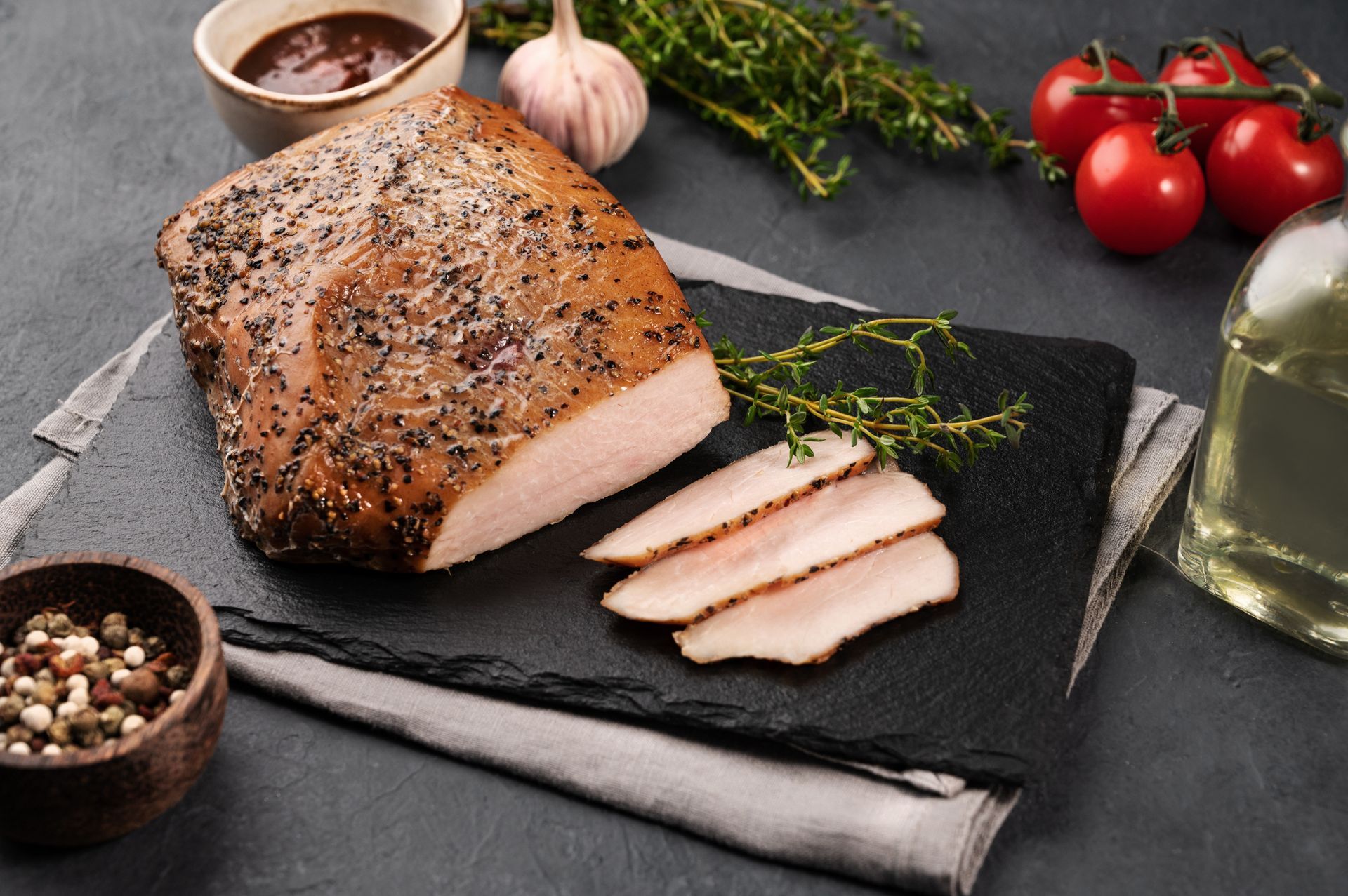 Smoked meat with sliced portions on a slate with thyme, garlic, tomatoes, and peppercorns.