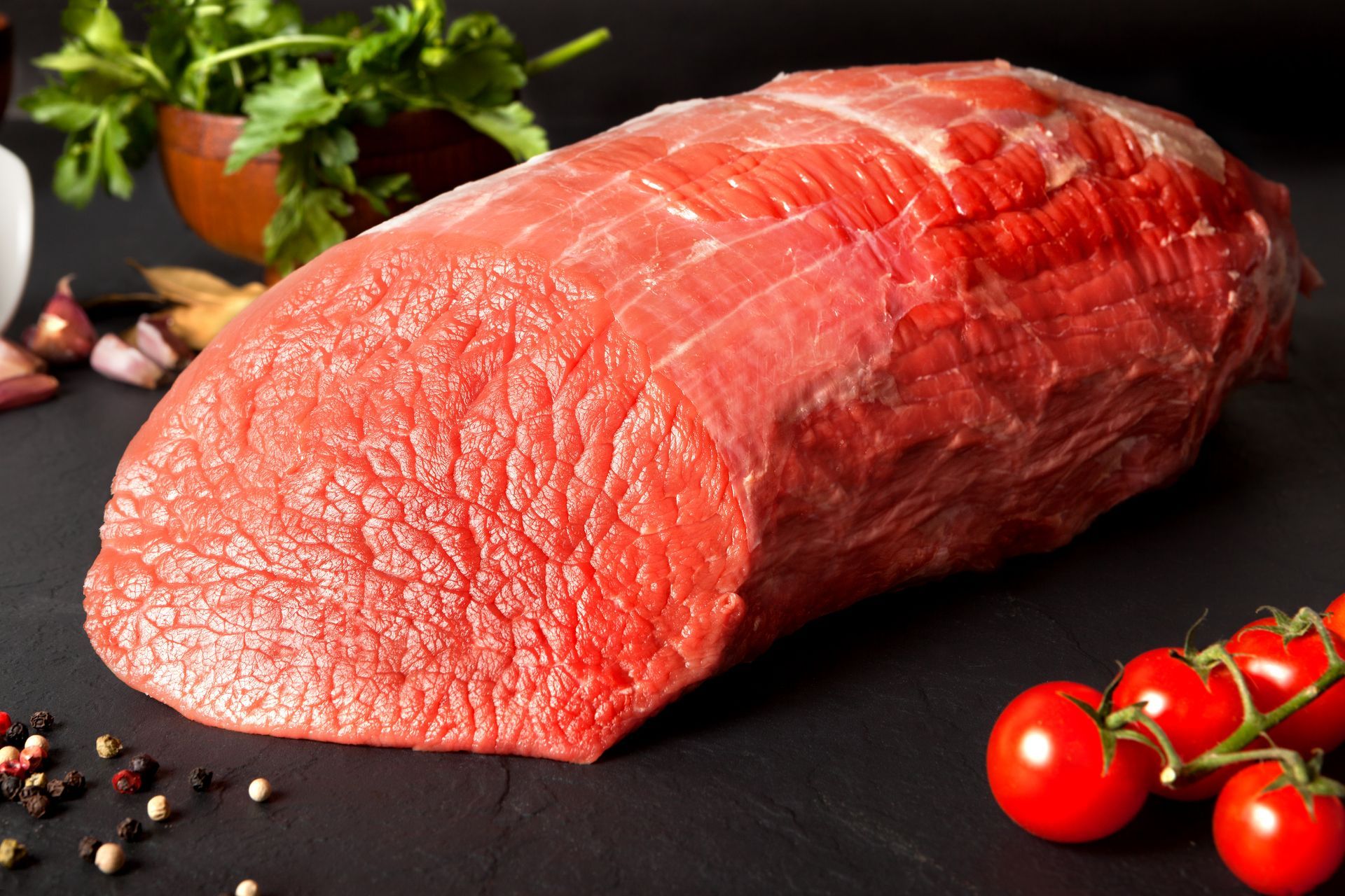 Raw beef roast, red with visible grain, surrounded by tomatoes, herbs, and spices on a black surface.