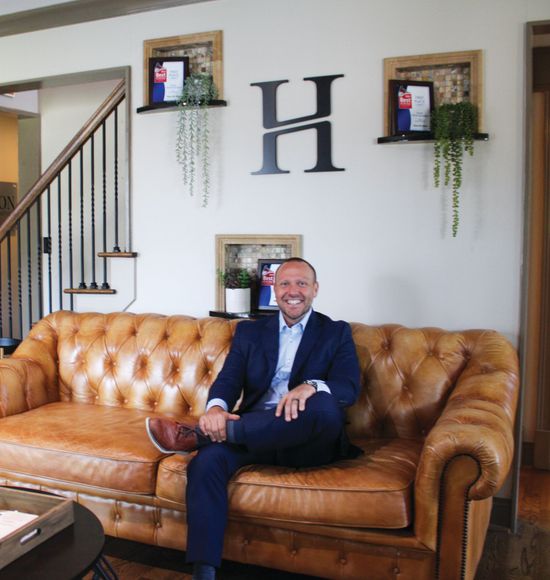 A smiling person in a blue suit sits on a tufted tan leather couch in front of a wall with a large letter H logo.