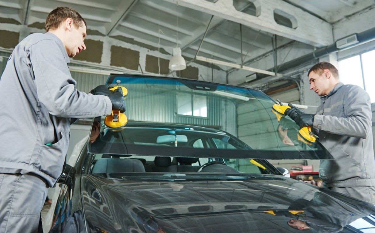 Two mechanics in gray uniforms working on a car windshield in a garage