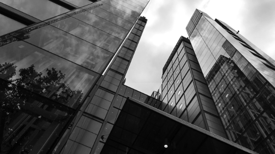 Fotografia em preto e branco de edifícios altos com janelas de vidro refletivo inclinadas para cima, em direção ao céu.