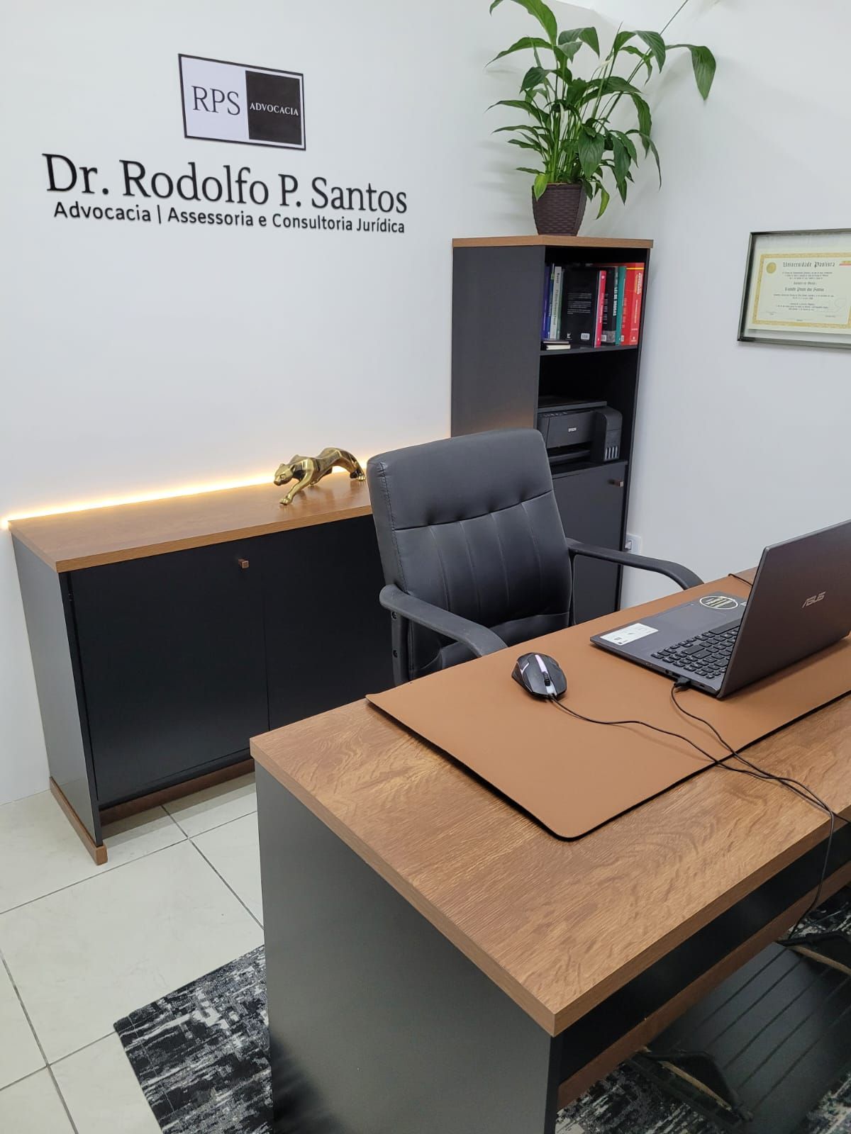 Escritório com mesa, computador, cadeira preta, prateleiras e placa com o nome do Dr. Rodolfo P. Santos, Advogado.