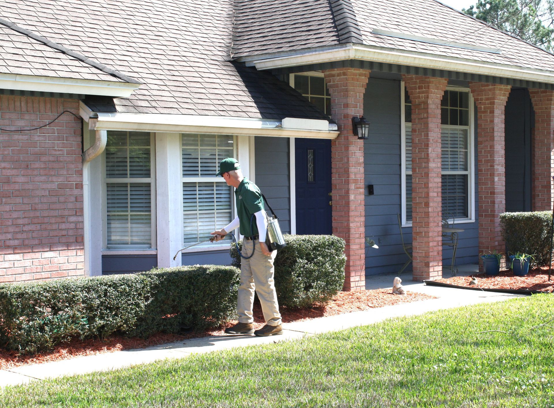 Mail carrier in green uniform delivers mail to a suburban house with brick and blue siding.