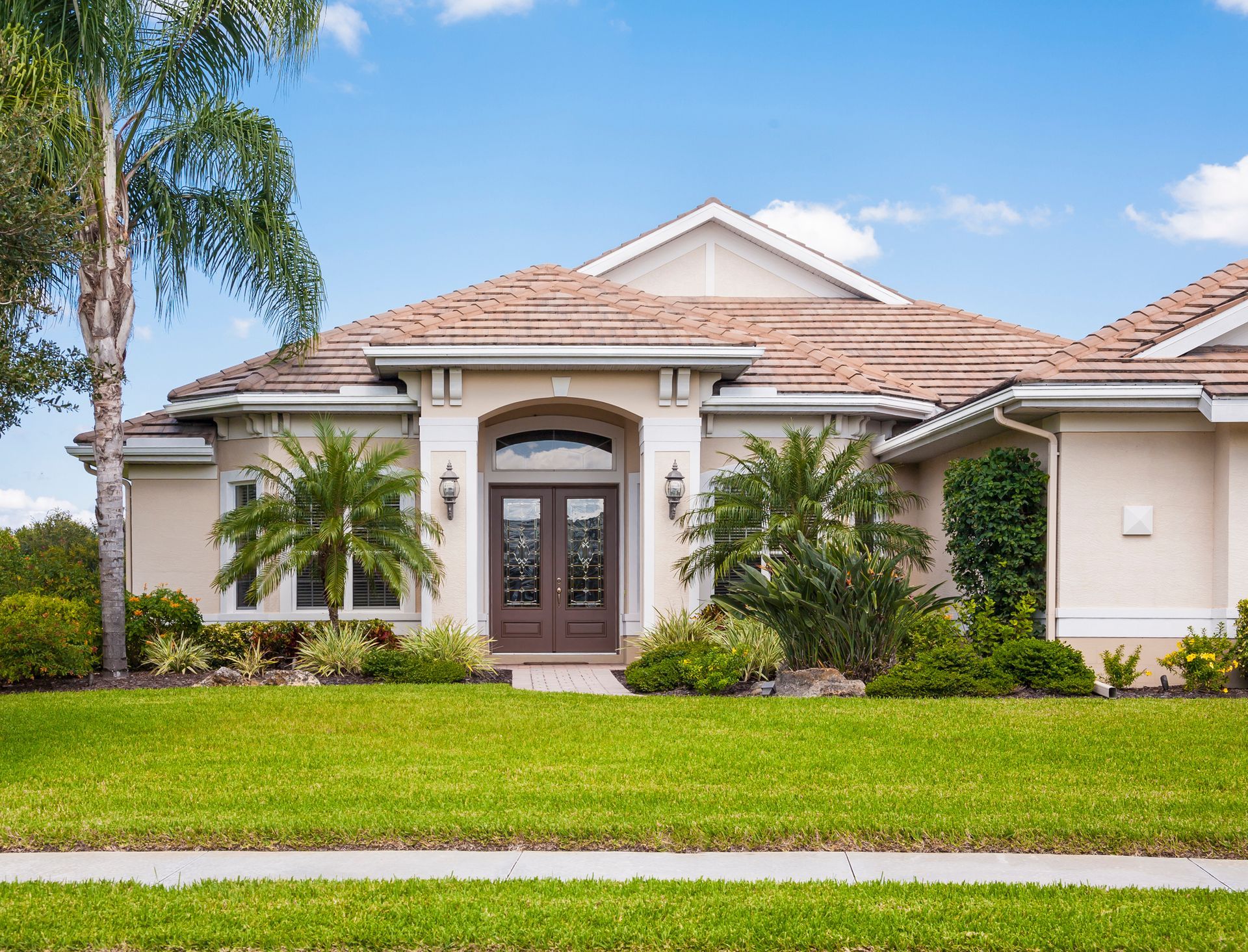 A contemporary home under a blue sky with palm trees, a green front lawn, and sidewalk.