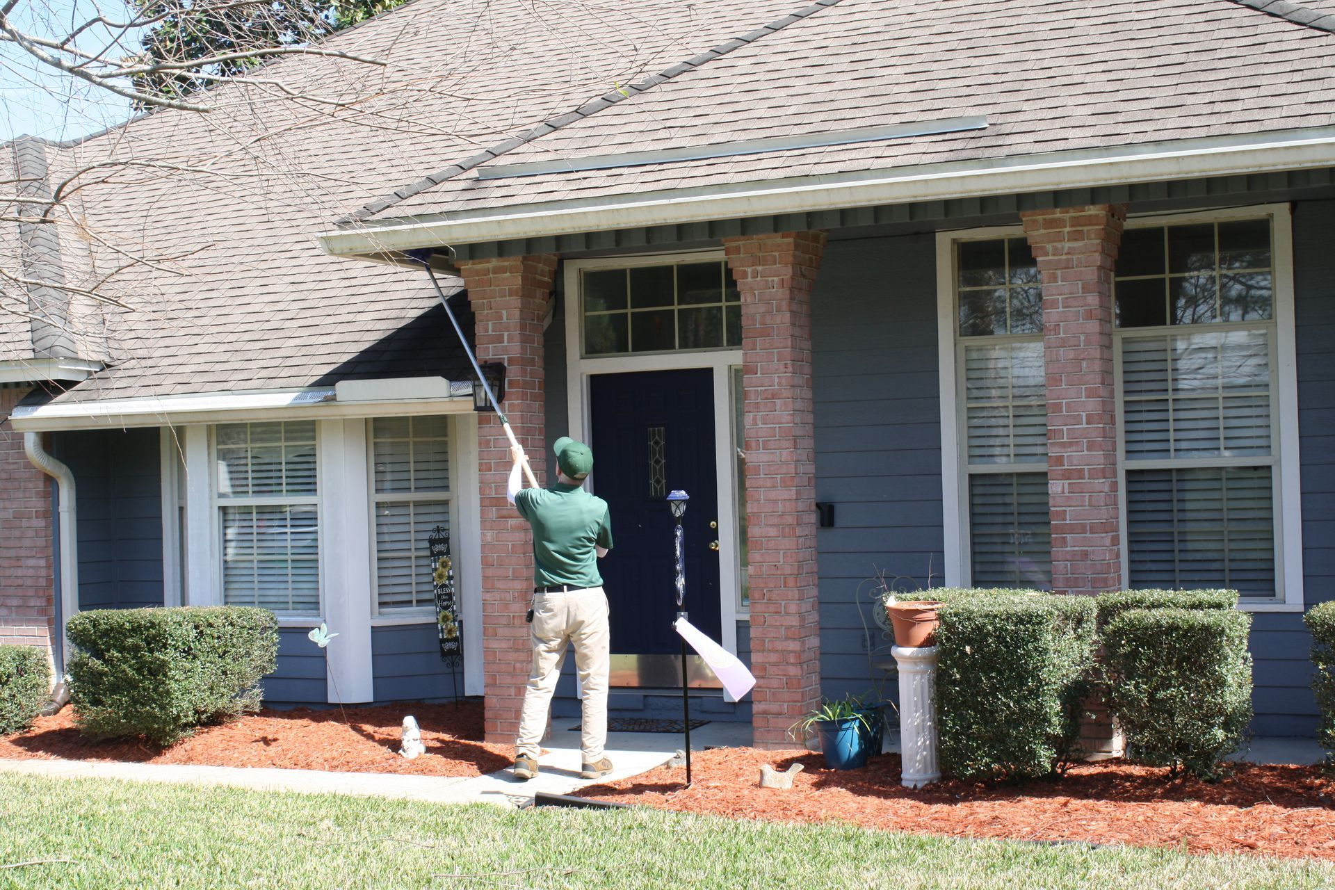 Person cleaning the front of a blue house with a long-handled tool. The house has brown roof and red mulch.