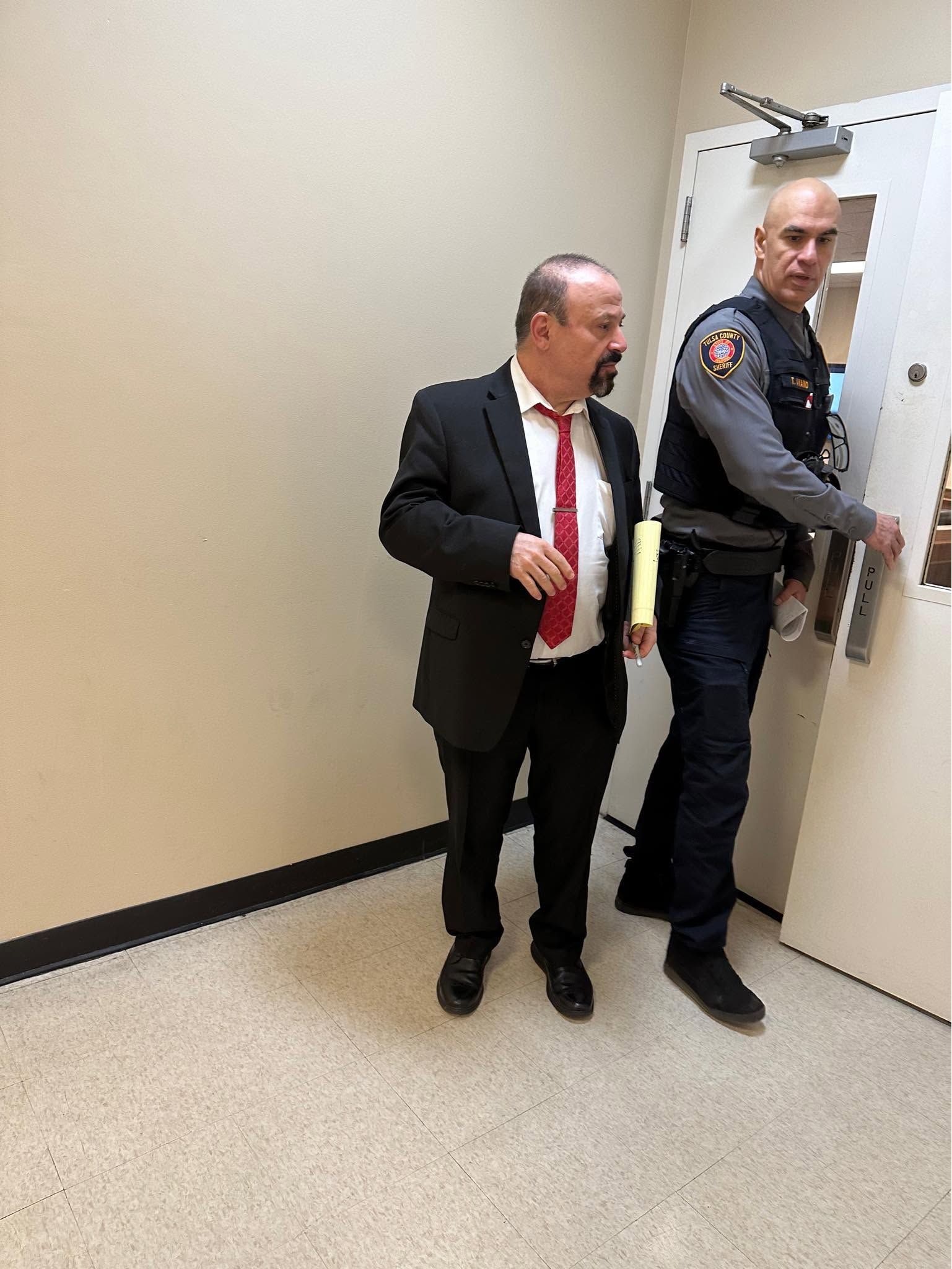 Two men stand by a hallway door, one in a suit and one in a security uniform opening it.