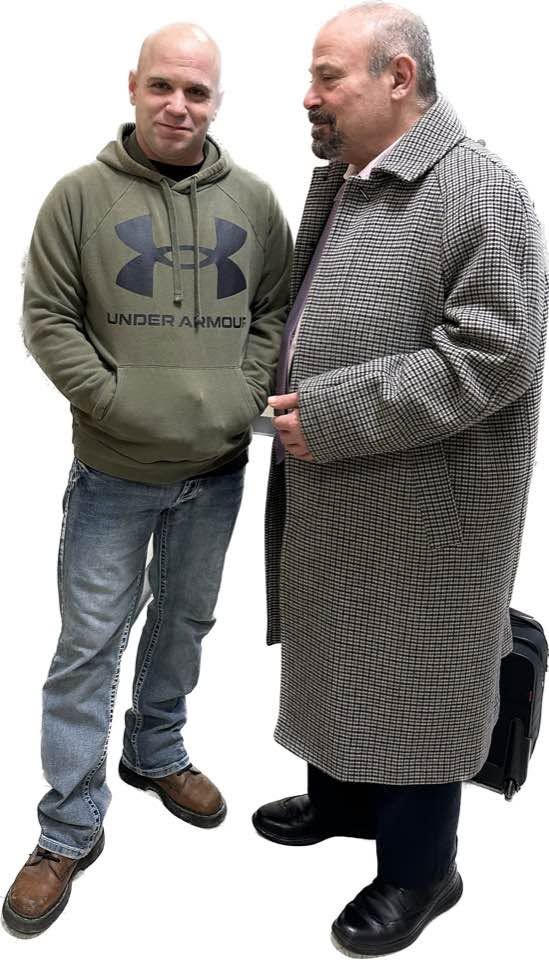 Two men standing together; one in a green hoodie and jeans, the other in a long patterned coat. Two men standing together; one in a green hoodie and jeans, the other in a long patterned coat.