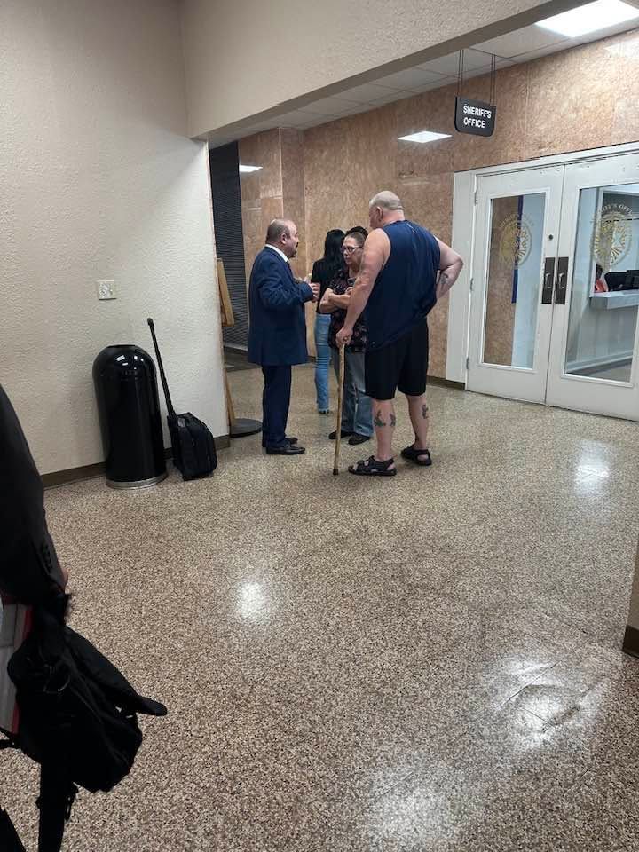 People standing and talking in a building lobby near glass doors and a wall-mounted sign.