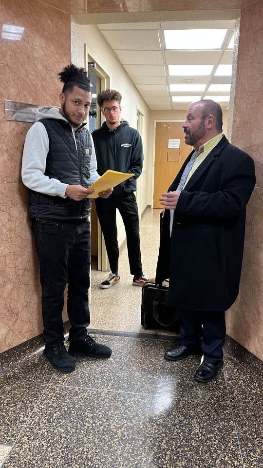 Three men standing in a hallway, one reading a yellow folder while another listens.