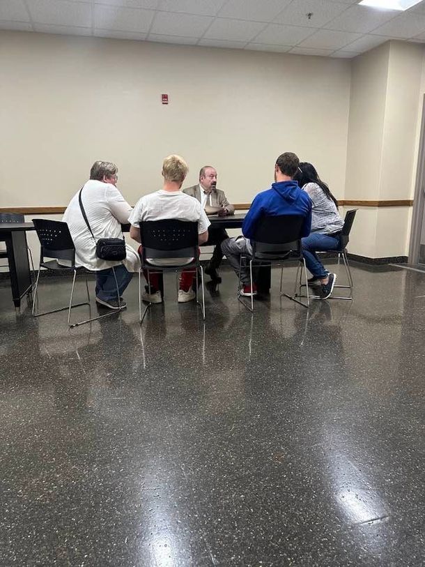 Five people seated in a circle at a table in a plain meeting room with beige walls and a glossy black floor. Five people seated in a circle at a table in a plain meeting room with beige walls and a glossy black floor.