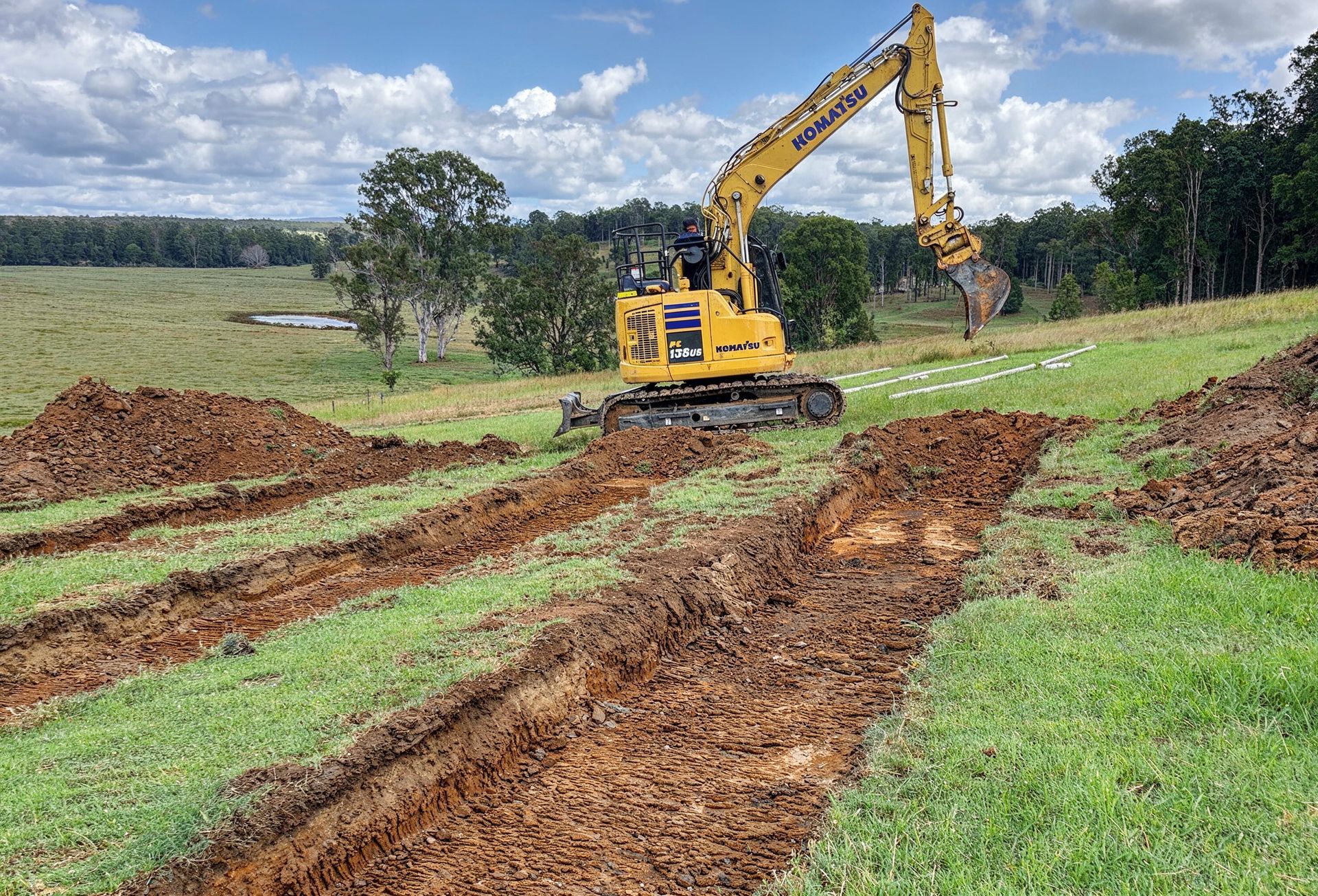 A yellow excavator digs trenches in a grassy field under a partly cloudy sky.