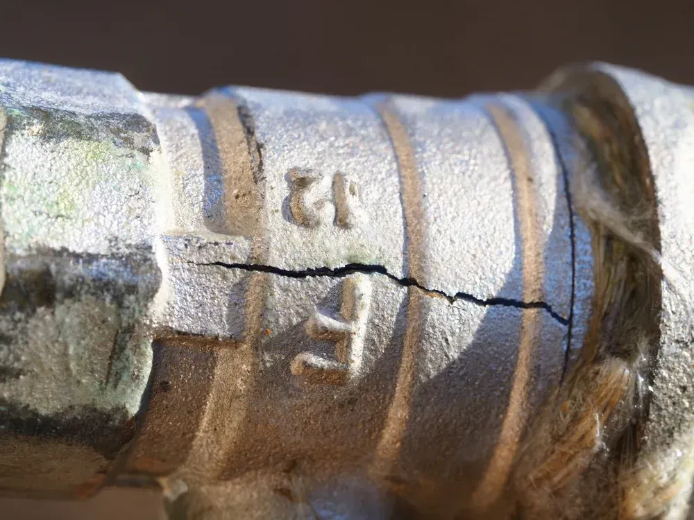 A close-up of a metal pipe fitting featuring a distinct, horizontal crack running through the cast material.