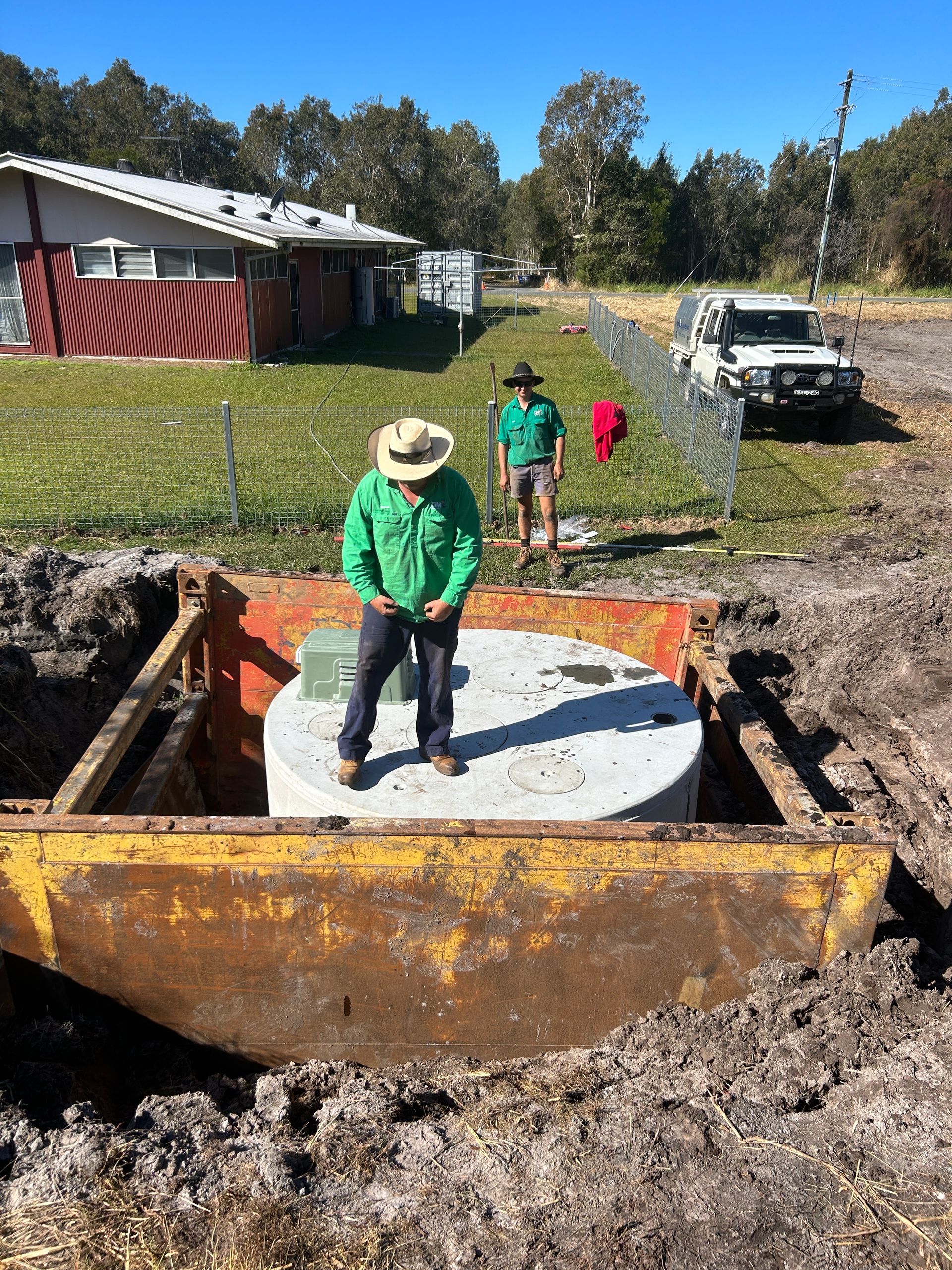 Two workers stand near a large circular concrete tank installed in an excavation pit, with a white truck parked nearby.