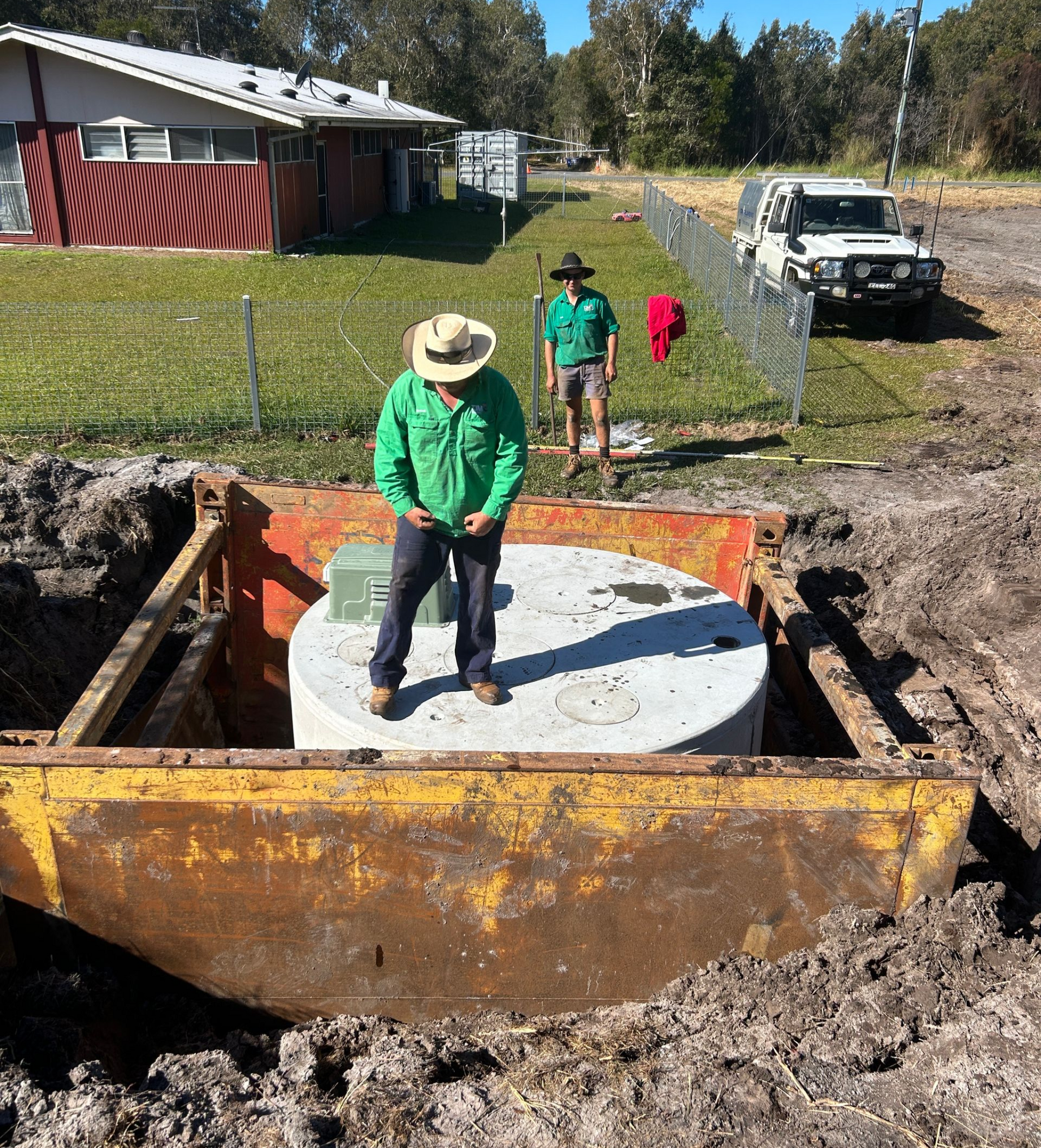 A person stands atop a concrete cylinder inside a trench box, while another person stands nearby on a grassy site.
