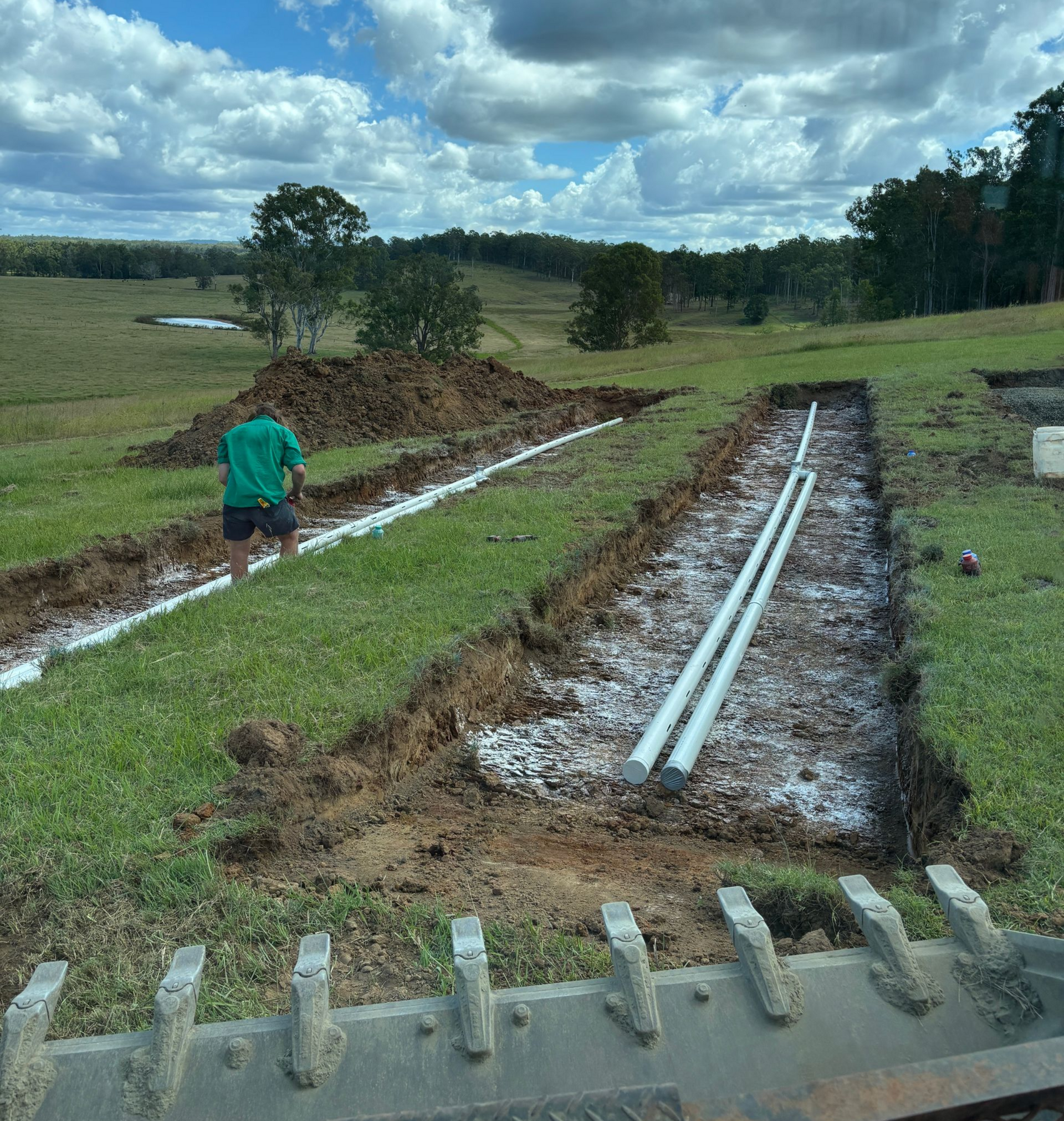 A person in a green shirt works on a trench with drainage pipes laid in crushed rock in a grassy rural landscape.