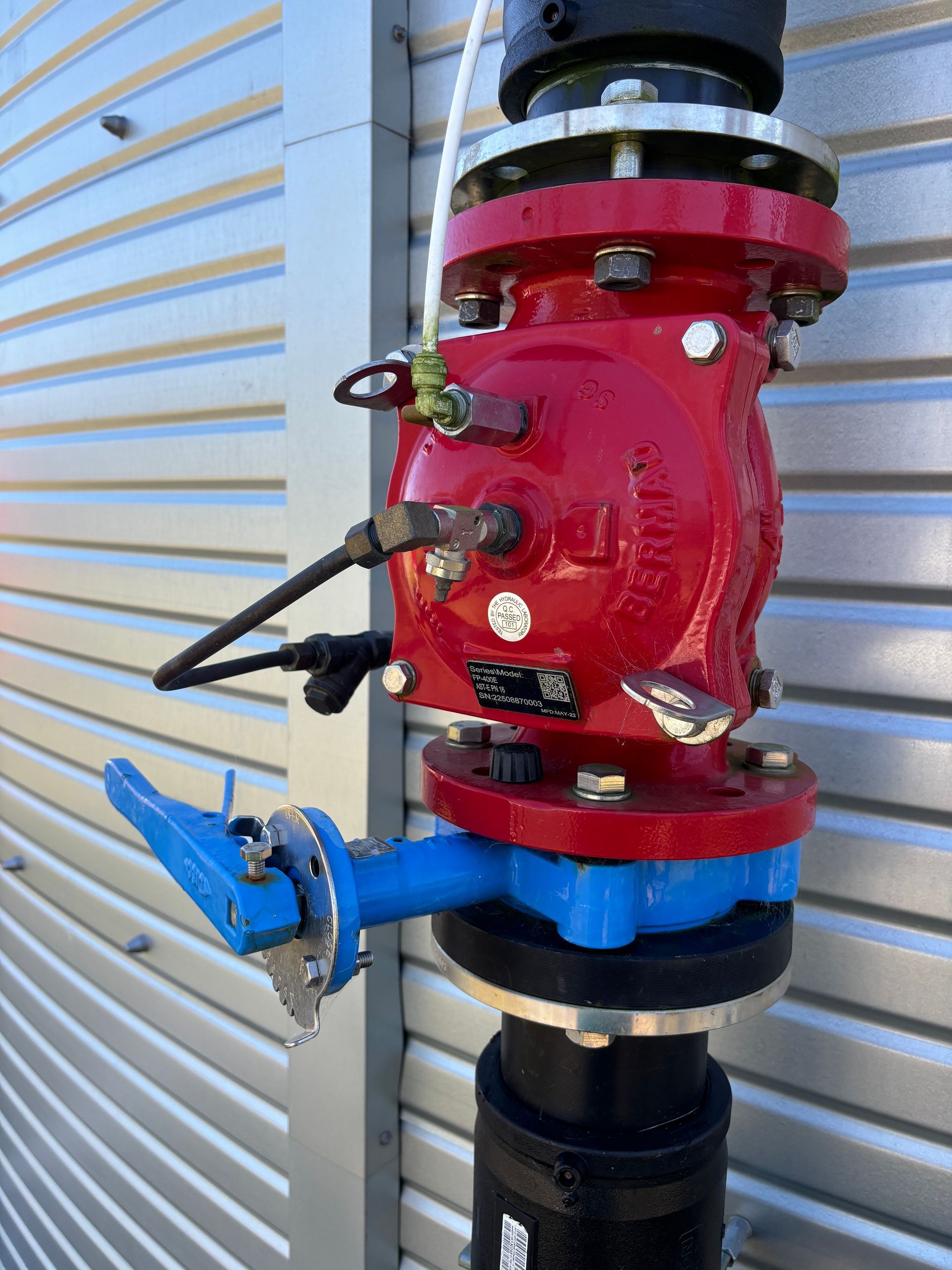 A red industrial valve with a blue side-lever handle mounted on a pipe against a corrugated metal wall.