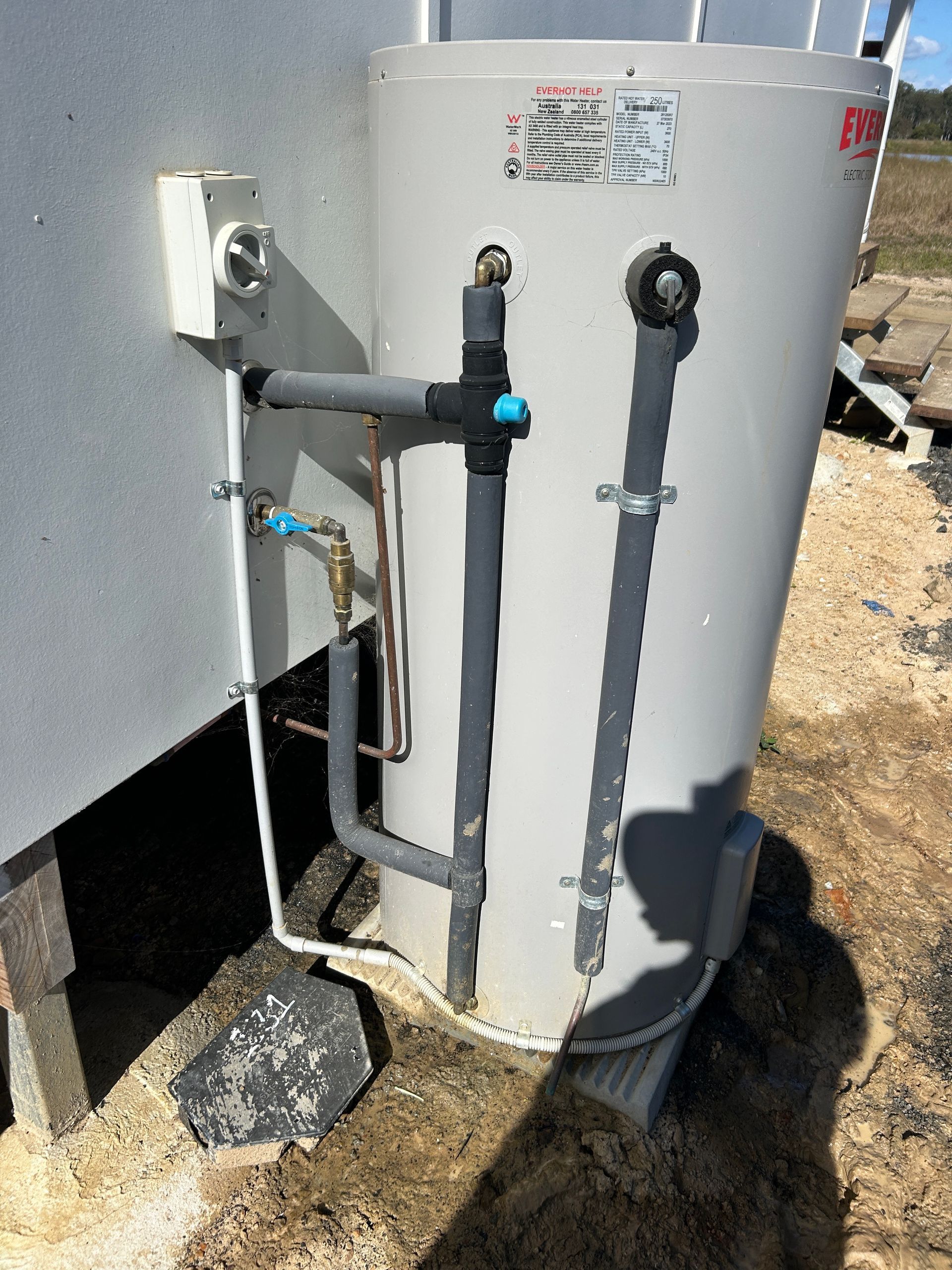 An outdoor electric water heater with grey insulated pipes and a disconnect box installed against a white exterior wall.