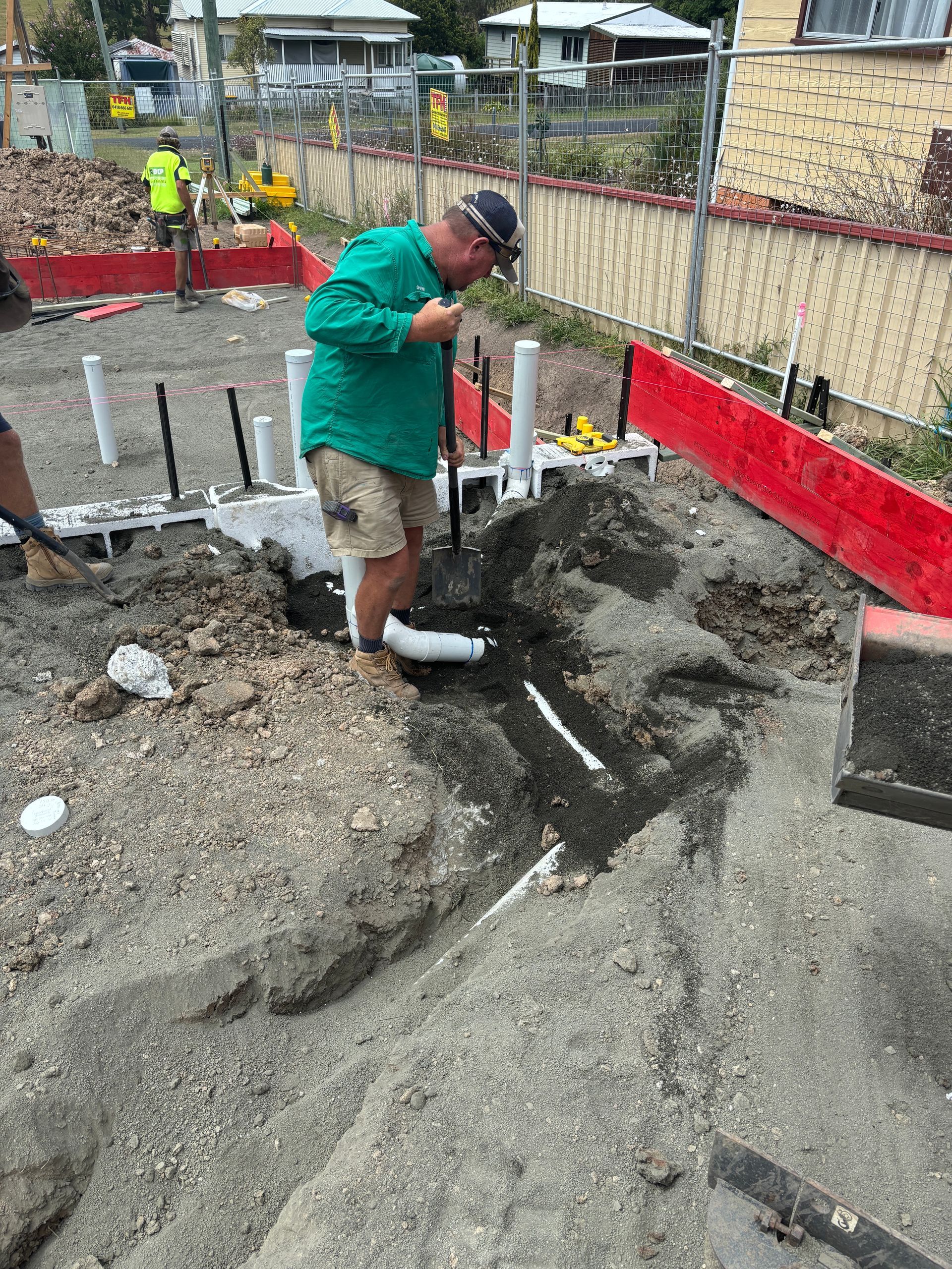 A construction worker in a green shirt digs a trench in the dirt of a residential building site.