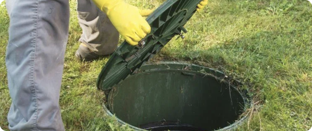 A person wearing yellow gloves lifting the green plastic cover of an outdoor septic or drainage tank in a grassy yard.
