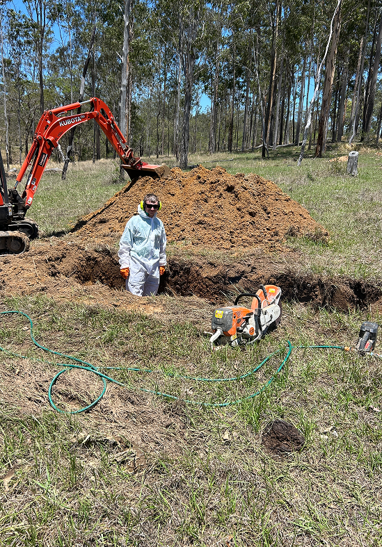 A person in white protective coveralls stands near an open trench and a mound of dirt next to an orange excavator.