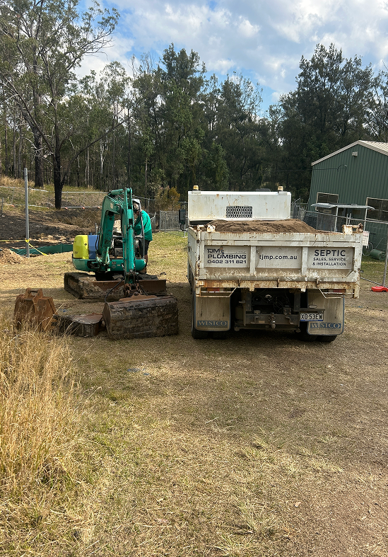 A teal excavator and a white dump truck parked on a dry, grassy plot near a shed and treeline.