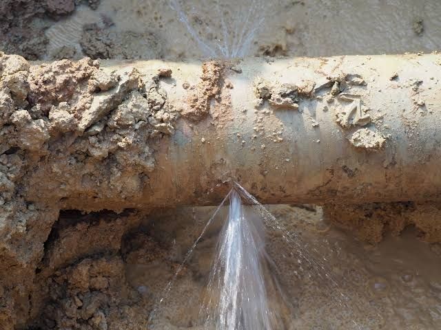 A buried metal pipe spraying water from multiple leaks into surrounding mud.