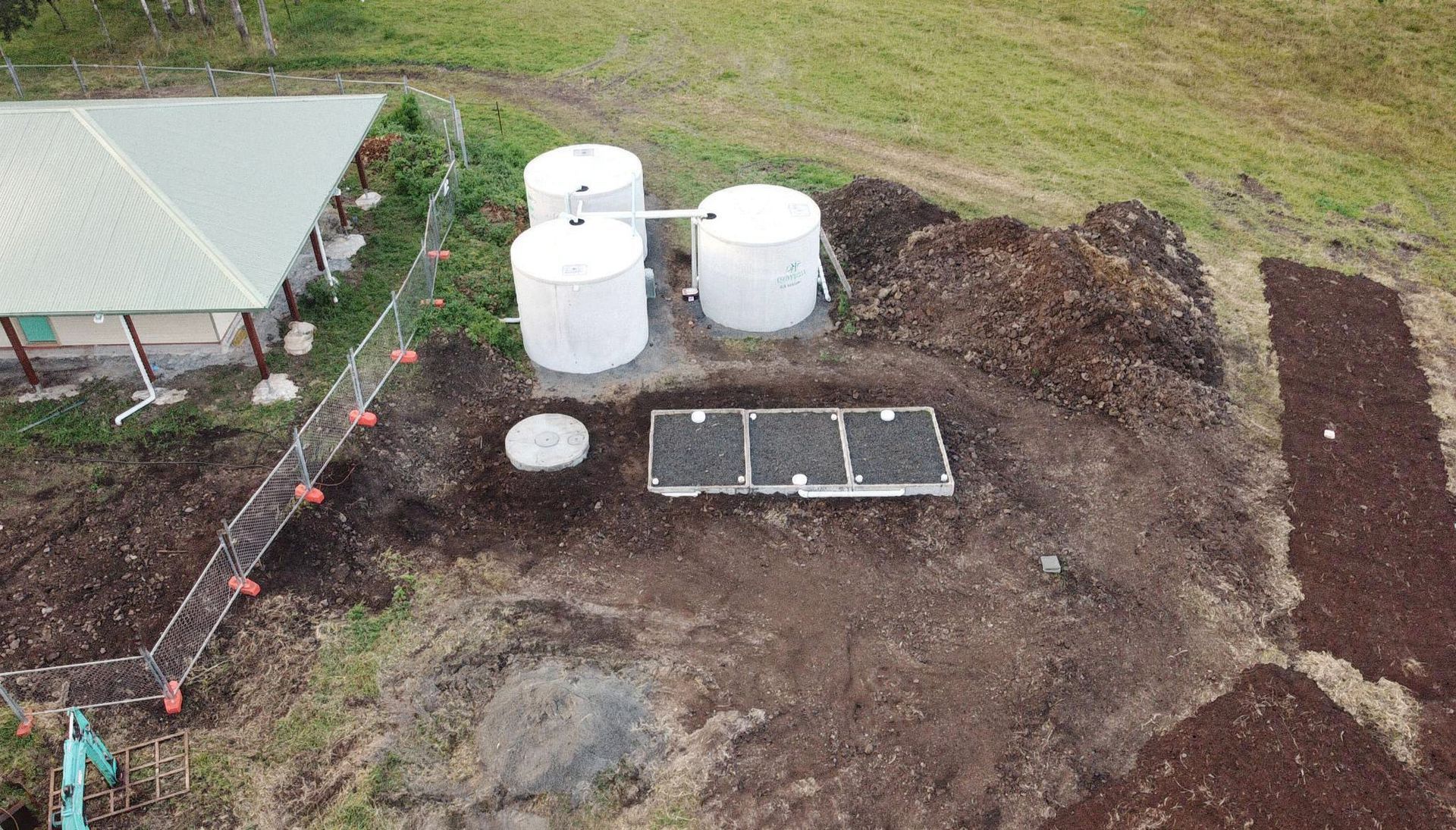 An aerial view shows three white cylindrical water tanks next to an in-ground septic system and landscaped garden beds.