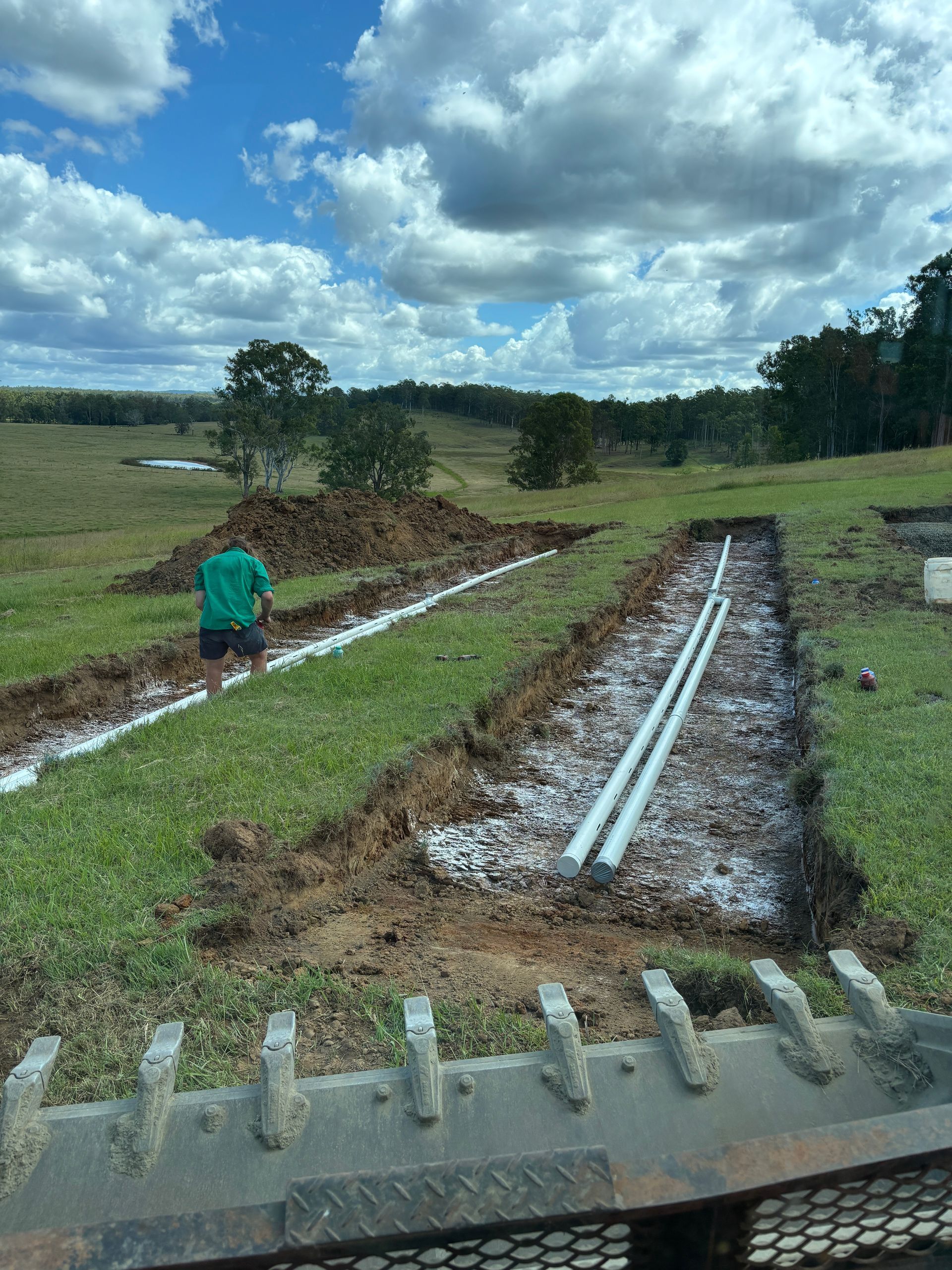 A person works on septic drain trenches with white PVC pipes laid in gravel-filled channels in a grassy field.