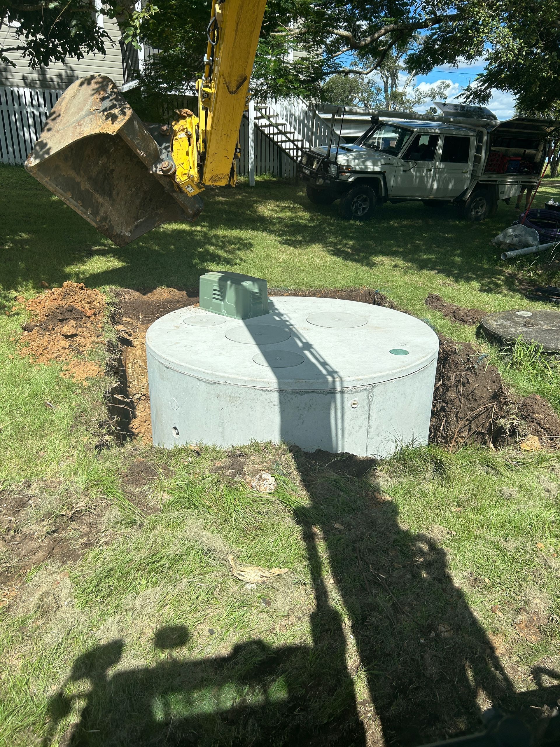 An excavator bucket lowers a large, circular concrete septic tank into a hole in a residential yard.